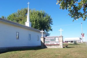 White country church with a steeple standing against the blue sky and bright green tree.