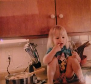 My daughter is sitting on the kitchen counter, licking her fingers. You can see the mixer in the background where we have mixed up cookie dough.