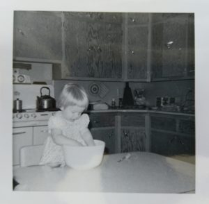 An old farmhouse kitchen with a small child standing on the chair at the table, with both hands in the cookie bowl.