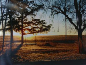 An old homemade swing set in the big backyard of a farm. The sun is setting in the background.