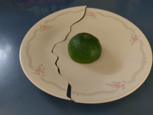 This picture is of a small white dessert plate with blue lines and clusters of small pink roses running around the edge. The plate sits on a blue counter, and has half of a lime upside down in its center.