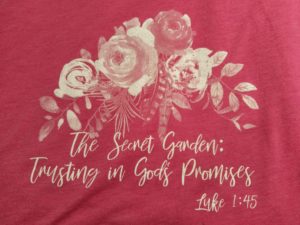 Pink T-shirt with white roses. Imprinted on the front is: "The Secret Garden: Trusting in God's Promises Luke 1:45"